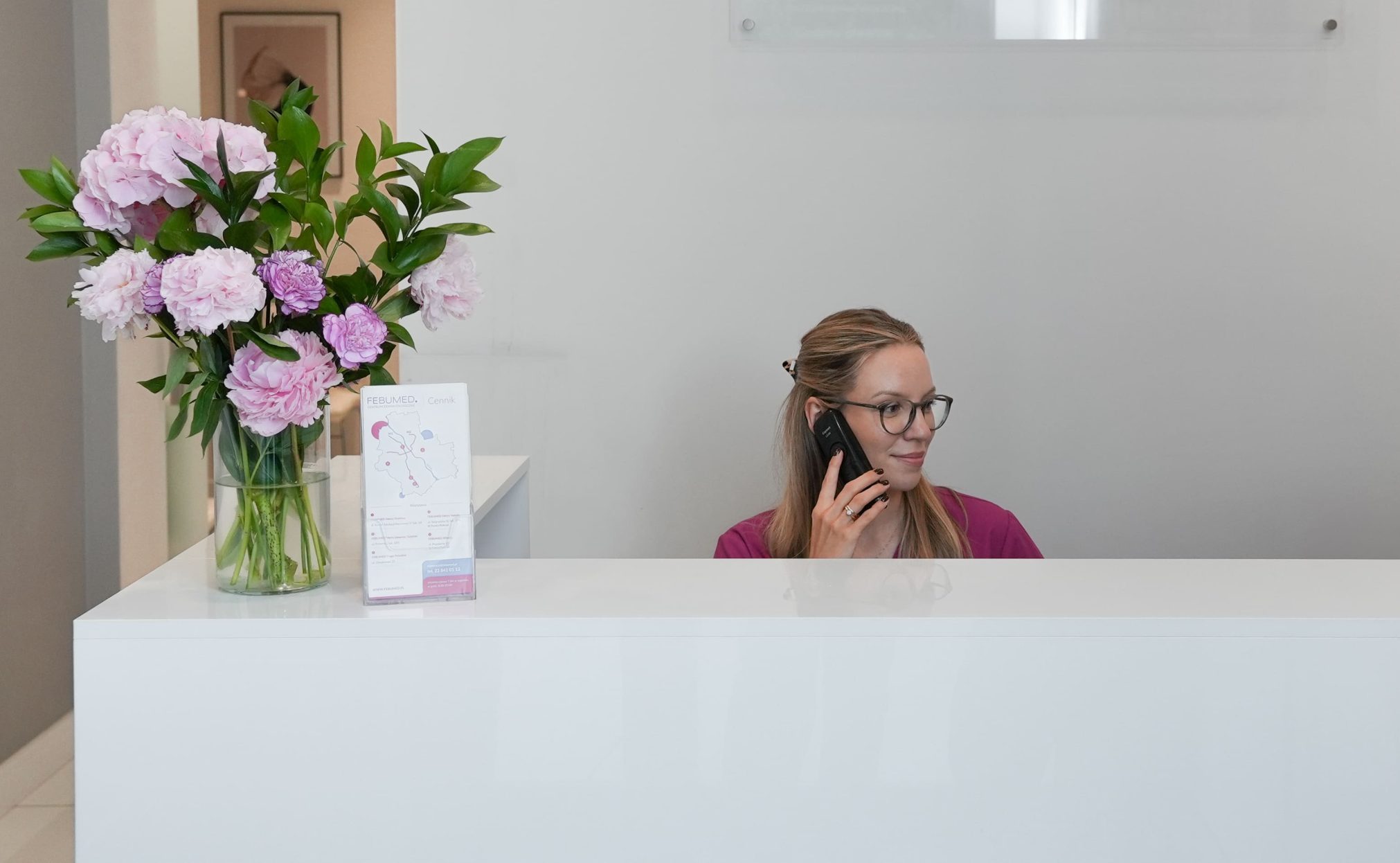 Recepcja Centrum Dermatologicznego FEBUMED w Warszawie - rejestratorka odbiera telefon, kwiaty na ladzie, w tle logotyp FEBUMED.
