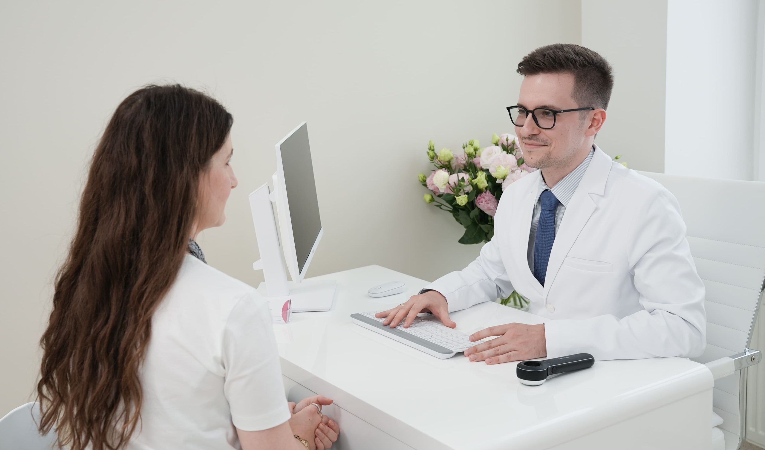 Uśmiechnięty doktor Rafał Studniarz w krawacie i białym fartuchu konsultuje pacjentkę w swoim gabinecie