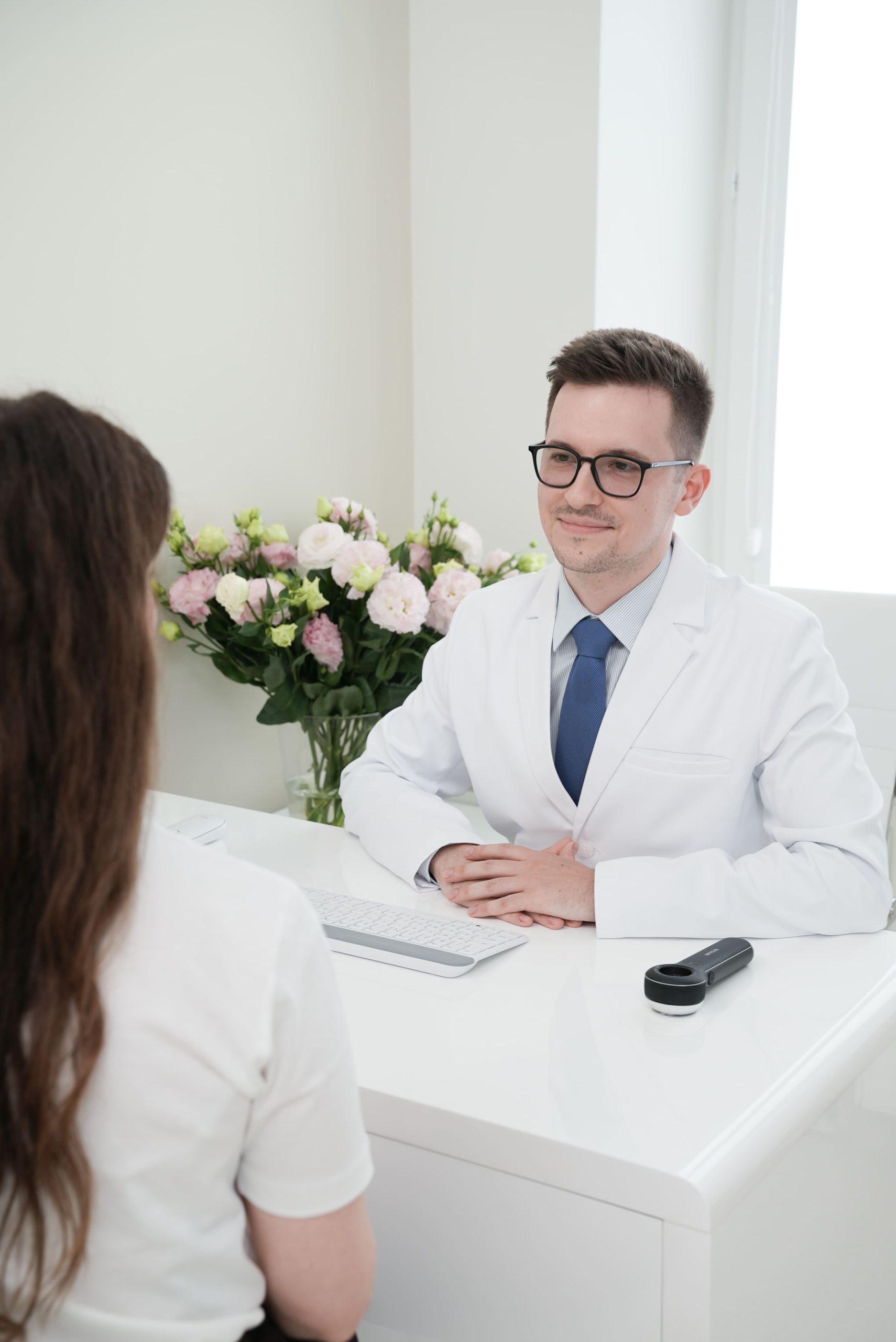 Uśmiechnięty doktor Rafał Studniarz w krawacie i białym fartuchu konsultuje pacjentkę w swoim gabinecie