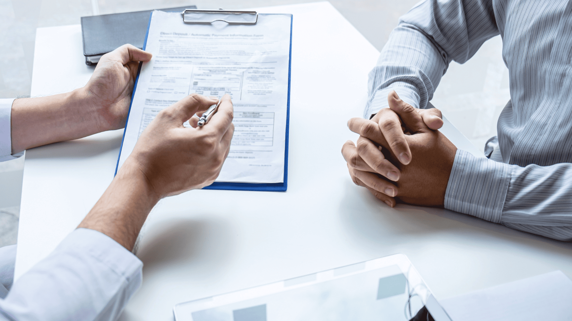 Dwie osoby siedzące przy białym stole. Osoba po lewej trzyma podkładkę z dokumentem i wskazuje coś na nim długopisem, podczas gdy osoba po prawej ma splecione dłonie i uważnie słucha. Obie osoby mają na sobie koszule, a w tle widać rozmyty fragment gabinetu.