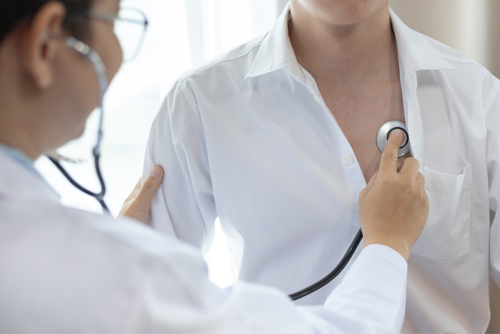 lekarz w fartuchu i w okularach bada stan płuc pacjenta ubranego w białą koszule za pomocą przykładania stetoskopu do klatki piersiowej.