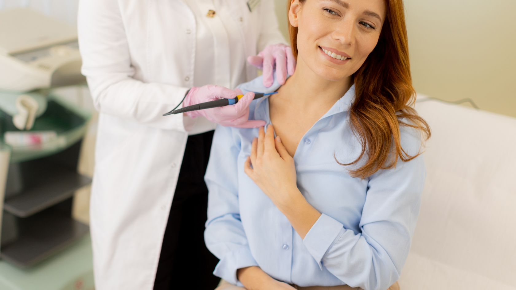 Stojący specjalista w fartuchu i różowych rękawiczkach medycznych usuwa zmiane skórną siedzącej rudej kobiecie w niebieskiej koszuli za pomocą elektrody w formie igły.