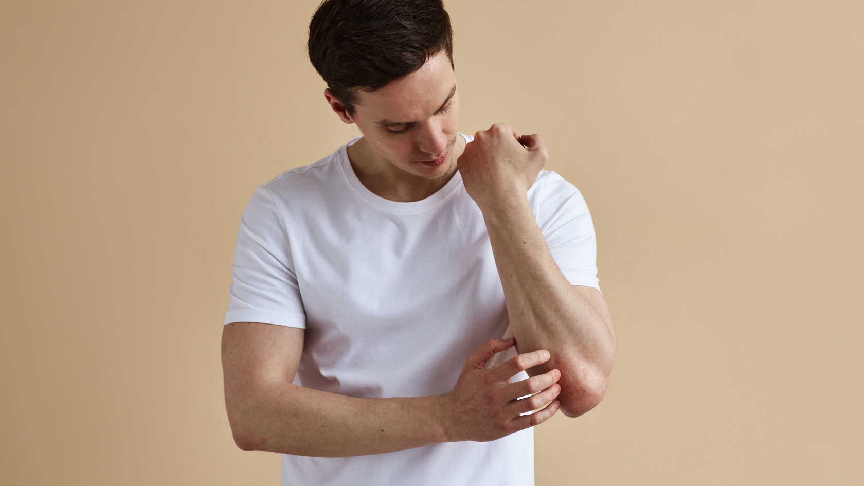 Młody mężczyzna w białej koszulce i krótko obciętymi, ciemnymi włosami, stojąc na żołtym tle drapie się po lewej ręce w okolicach łokcia. Patrzy na rękę, na której można zauważyć lekkie zaczerwienienie
