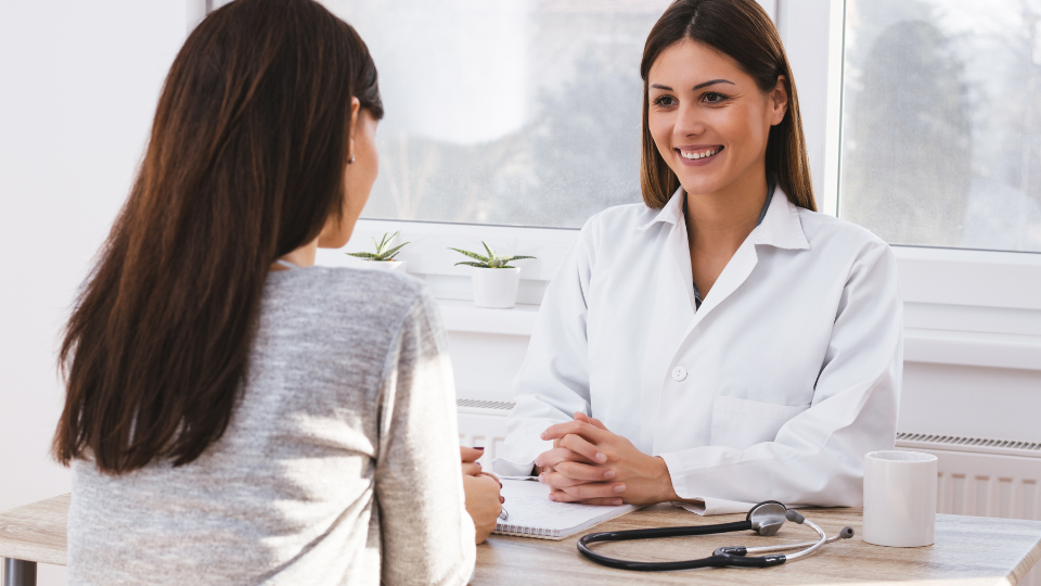 Uśmiechnięta młoda lekarka w białym fartuchu lekarskim rozmawiająca z siedzącą naprzeciwko niej pacjentką w szarej bluzce.