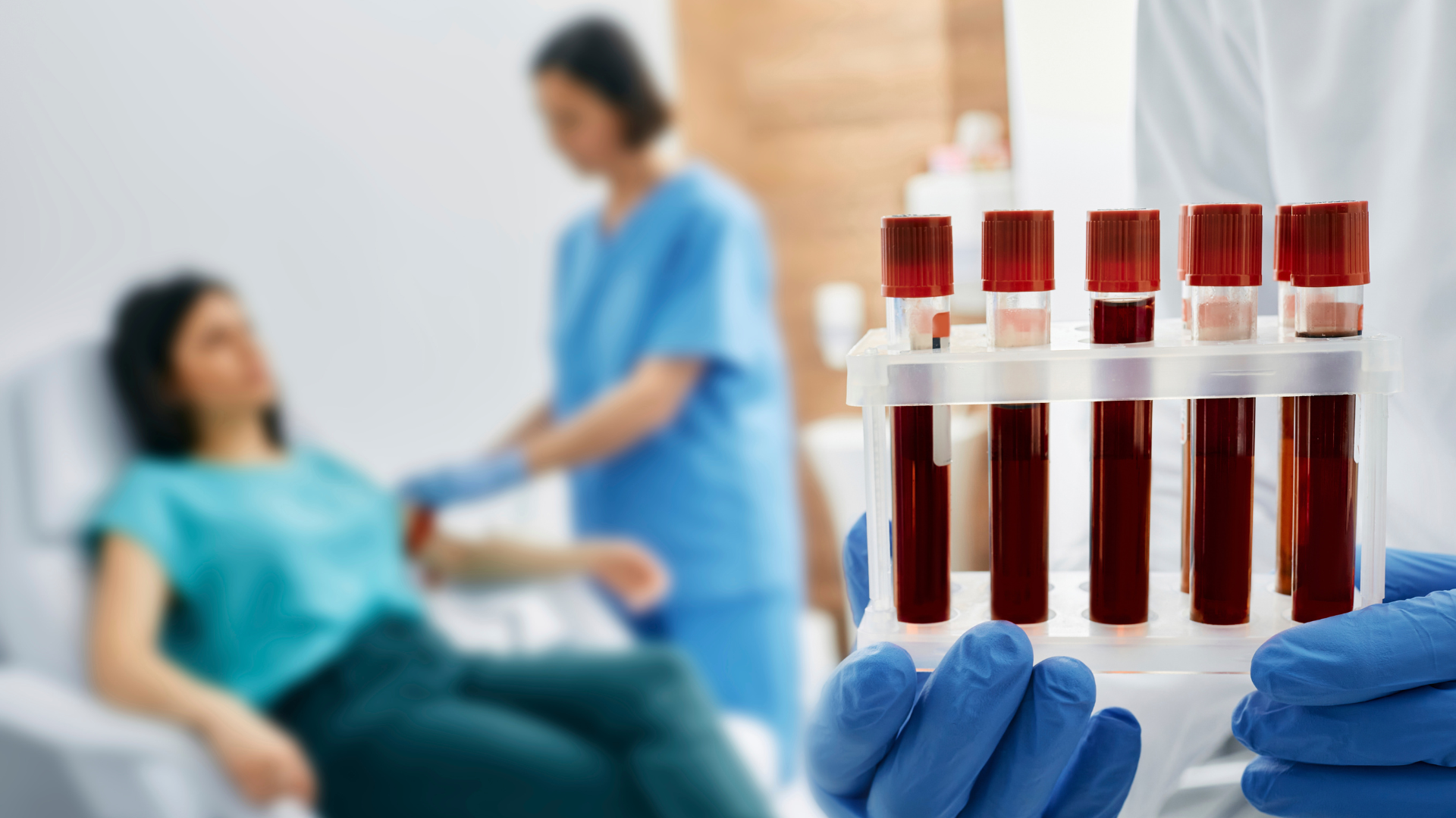 Lekarz w niebieskich rękawiczkach medycznych i białym fartuchu trzymający 5 próbek z krwią, w tle widać pielęgniarkę pobierającą krew leżącej pacjentce.