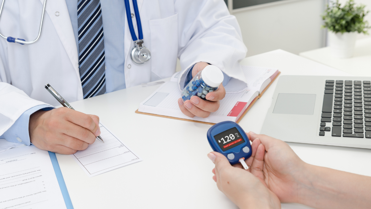 Lekarz diabetolog wydaje pacjentowi leki lewą ręką w tym samym czasie zapisując receptę prawą. Pacjent trzyma w rękach glukometr pokazujący wartość 128mg/dl.lekarz i pacjent siedzą przy biurku, w tle widać kwiatek oraz laptop lezacy na biurku.