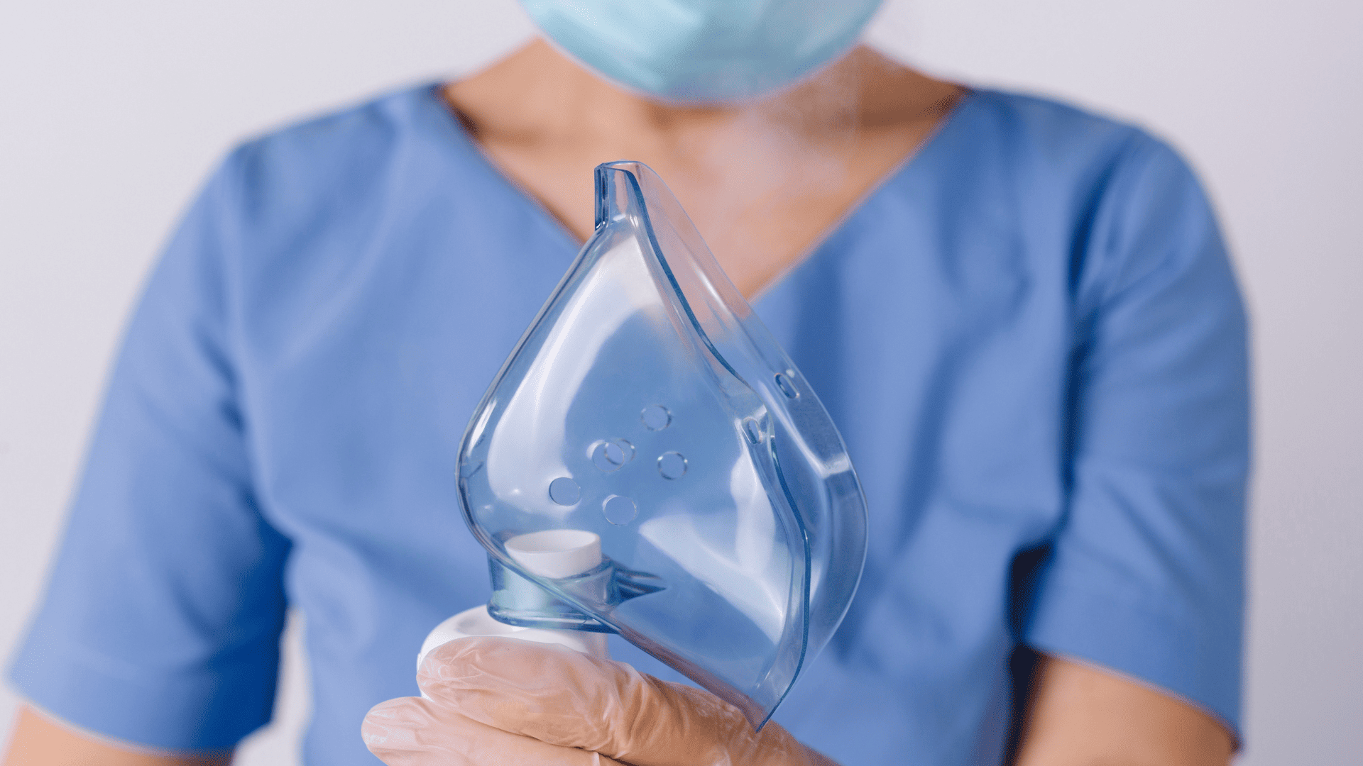 Pracownik medyczny w niebieskim stroju medycznym trzyma maskę do inhalacji w rękach na, których ma rękawiczki.