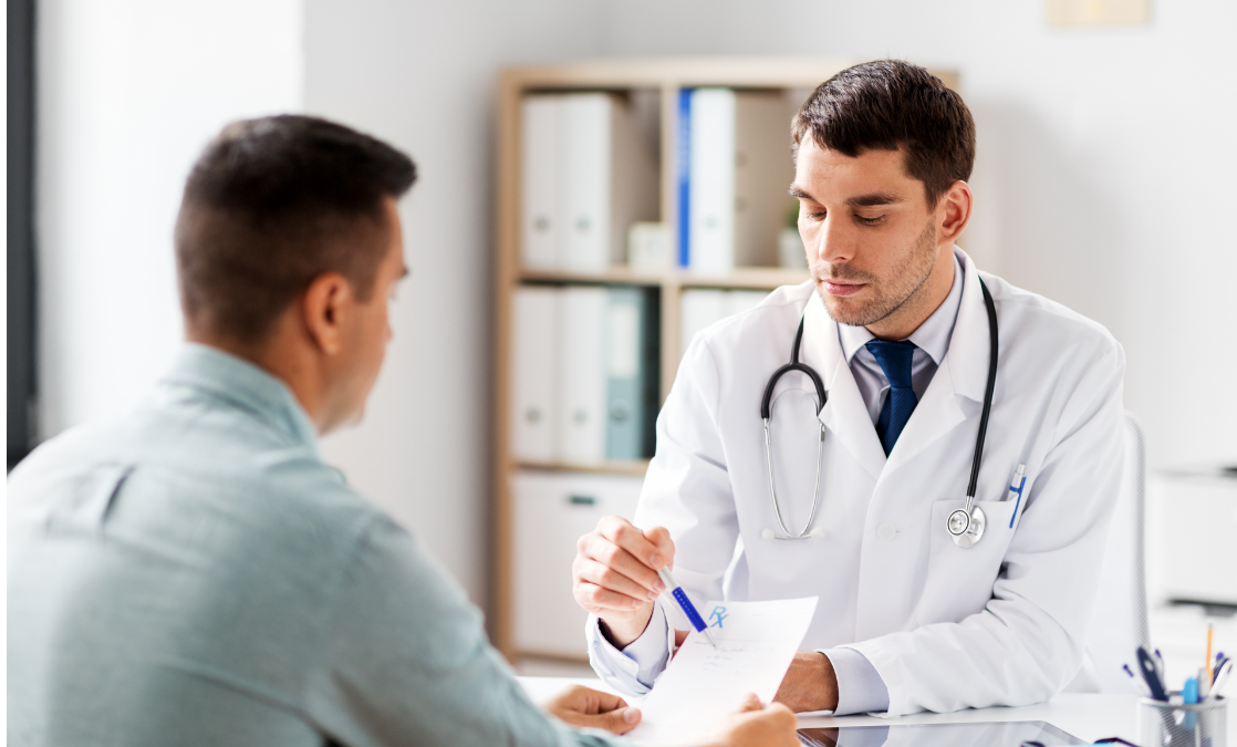 Lekarz w białym fartuchu ze stetoskopem założonym na szyje omawia i pokazuje pacjentowi na kartce wyniki badań, oboje siedzą przy biurku. W tle widać szafkę z segregatorami biurowymi.