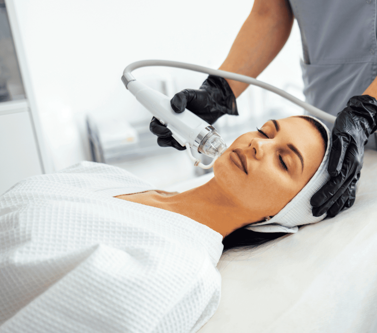 lekarz w czarnych rękawiczkach wykonująca zabieg radiofrekwencji mikroigłowej na twarzy leżącej na plecach pacjentki za pomocą białej głowicy urządzenia