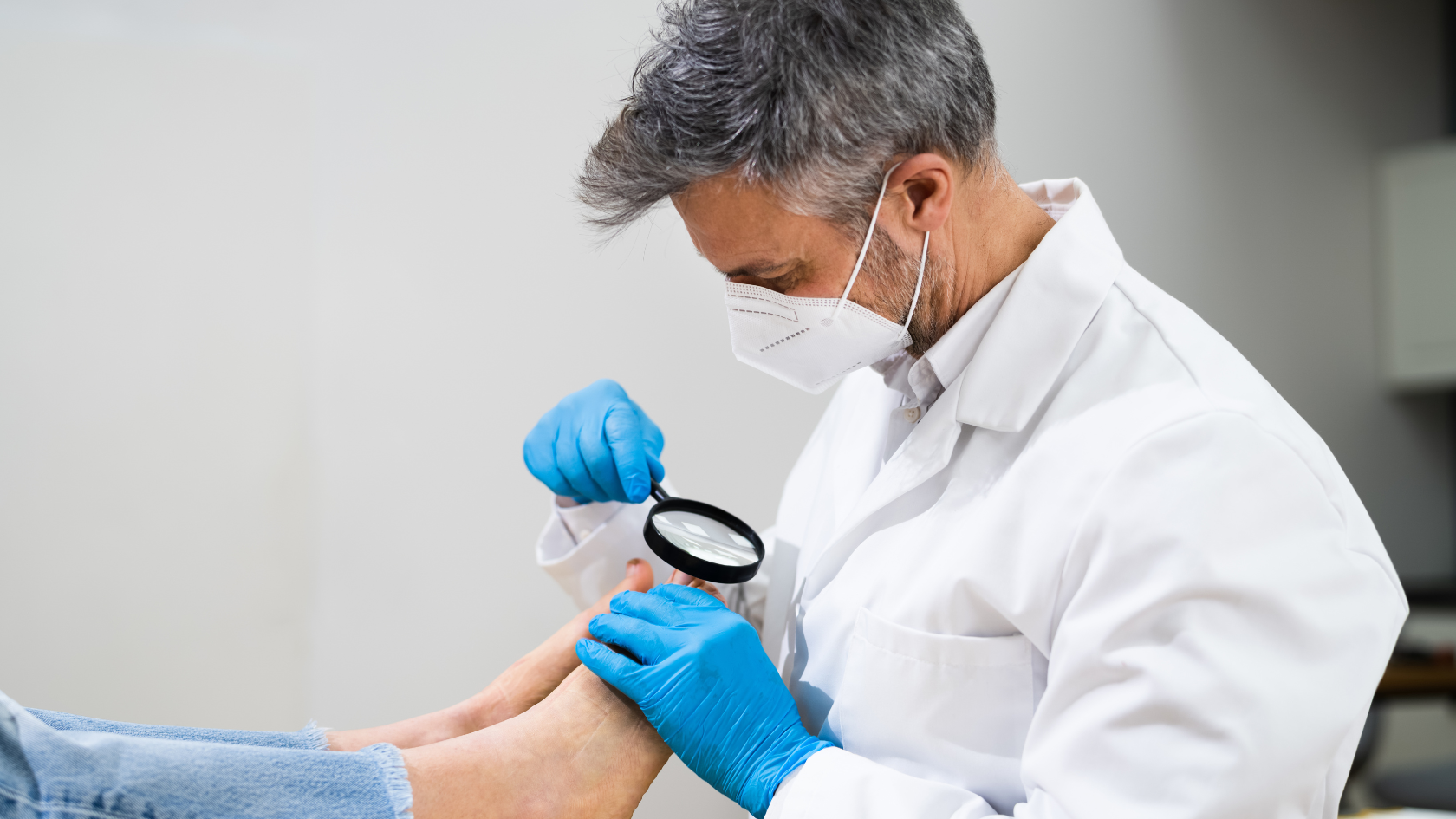 Chirurg w medycznych rękawiczkach, fartuchu i maseczce ochronnej kontrolujący stan paznokcia pacjenta leżącego na stole używając lupy.