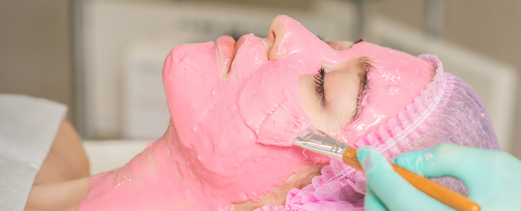 Kosmetolog wykonuje zabieg nawilżający twarz pacjentki, której skóra pokryta jest różowym preparatem