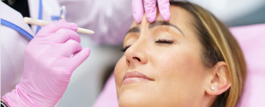 Lekarz w rękawiczkach przygotowuje pacjentkę do zabiegu medycyny estetycznej, zaznaczając miejsca aplikacji preparatu na czole