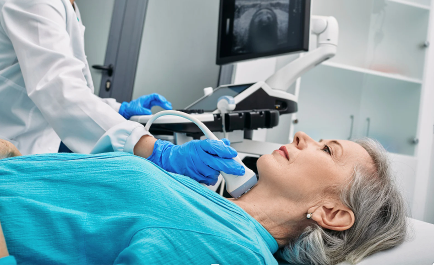 Starsza, siwa kobieta w niebieskiej bluzce i srebrnych kolczykach, leżąca na plecach podczas gdy badający ją lekarz w białym fartuchu i rękawiczkach medycznych bada jej szyje ultrasonografem. W tle widać telewizorek pokazujący obraz z ultrasonopgrafu