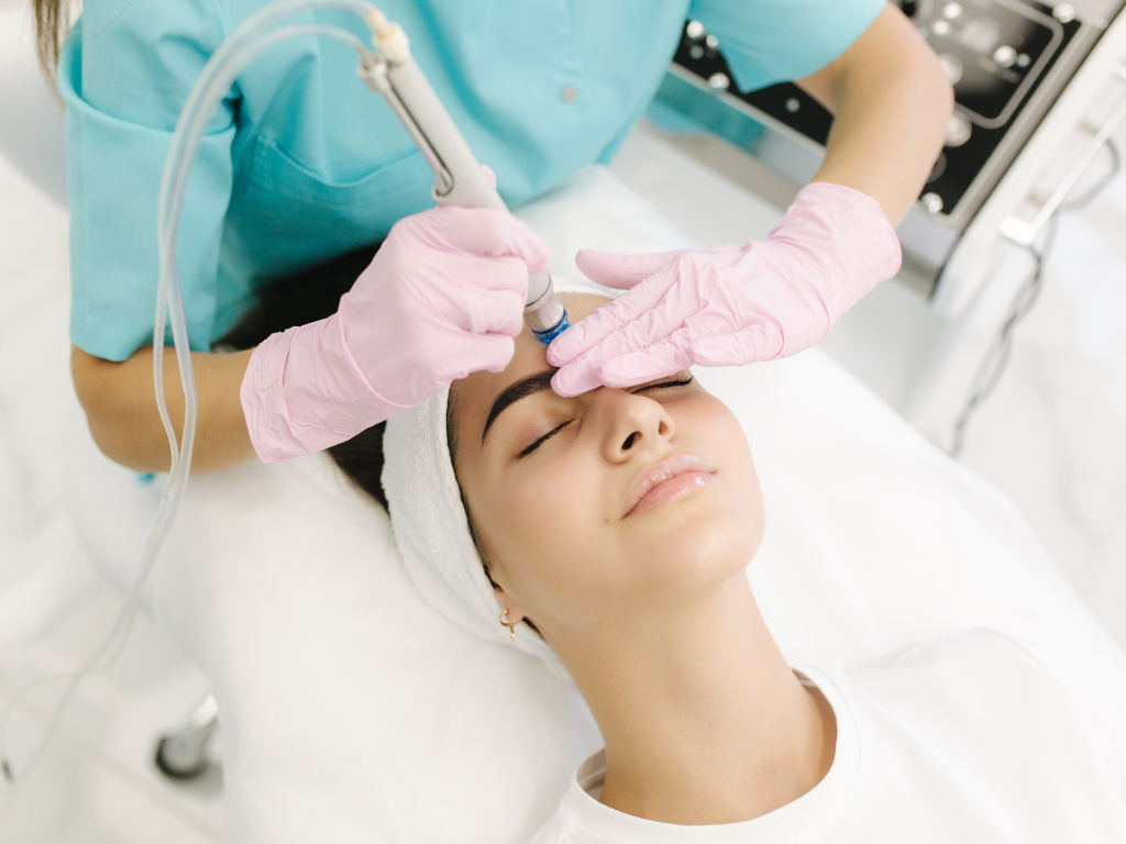 Kosmetolog w różowych rękawiczkach wykonuje oczyszczanie wodorowe na twarzy młodej pacjentki w gabinecie kosmetologicznym