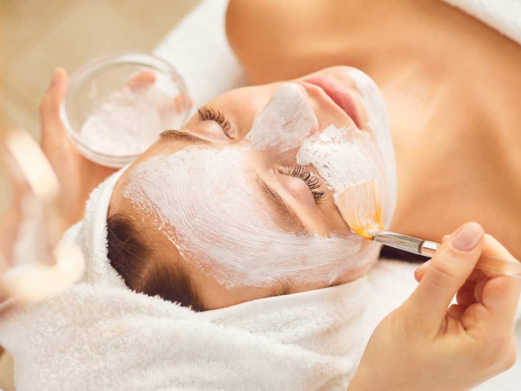 Kosmetolog nakłada biały preparat peelingu chemicznego na twarz pacjentki w gabinecie medycyny estetycznej