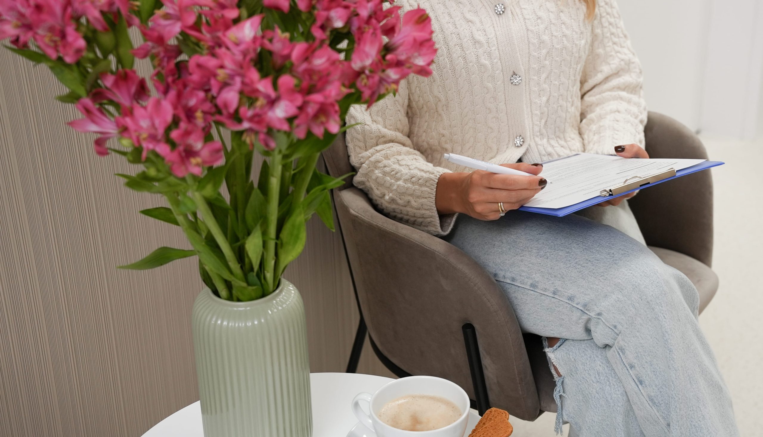 kobieta uzupełniająca formularz w poczekalni, na stoliku piękne różowe kwiaty i kawa z ciasteczkiem