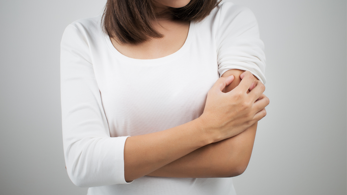 Młoda kobieta w białej bluzce i ciemnymi włosami, drapie się po lewym ramieniu na szarym tle.