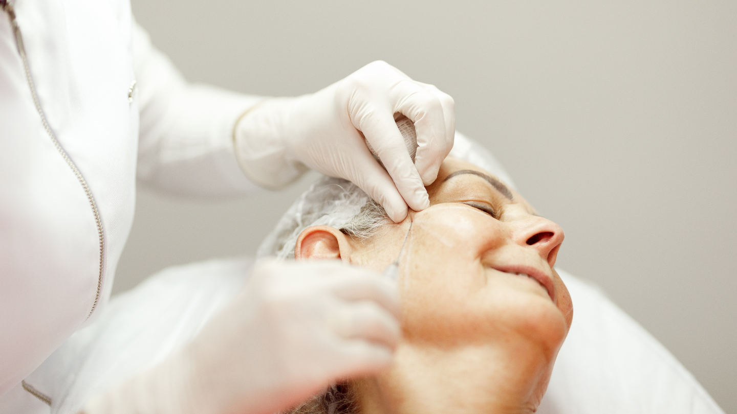 Lekarz w białym fartuchu i rękawiczkach medycznych precyzyjnie wbija igłę pod skórę twarzy starszej, uśmiechniętej pacjentki leżącej na stole.