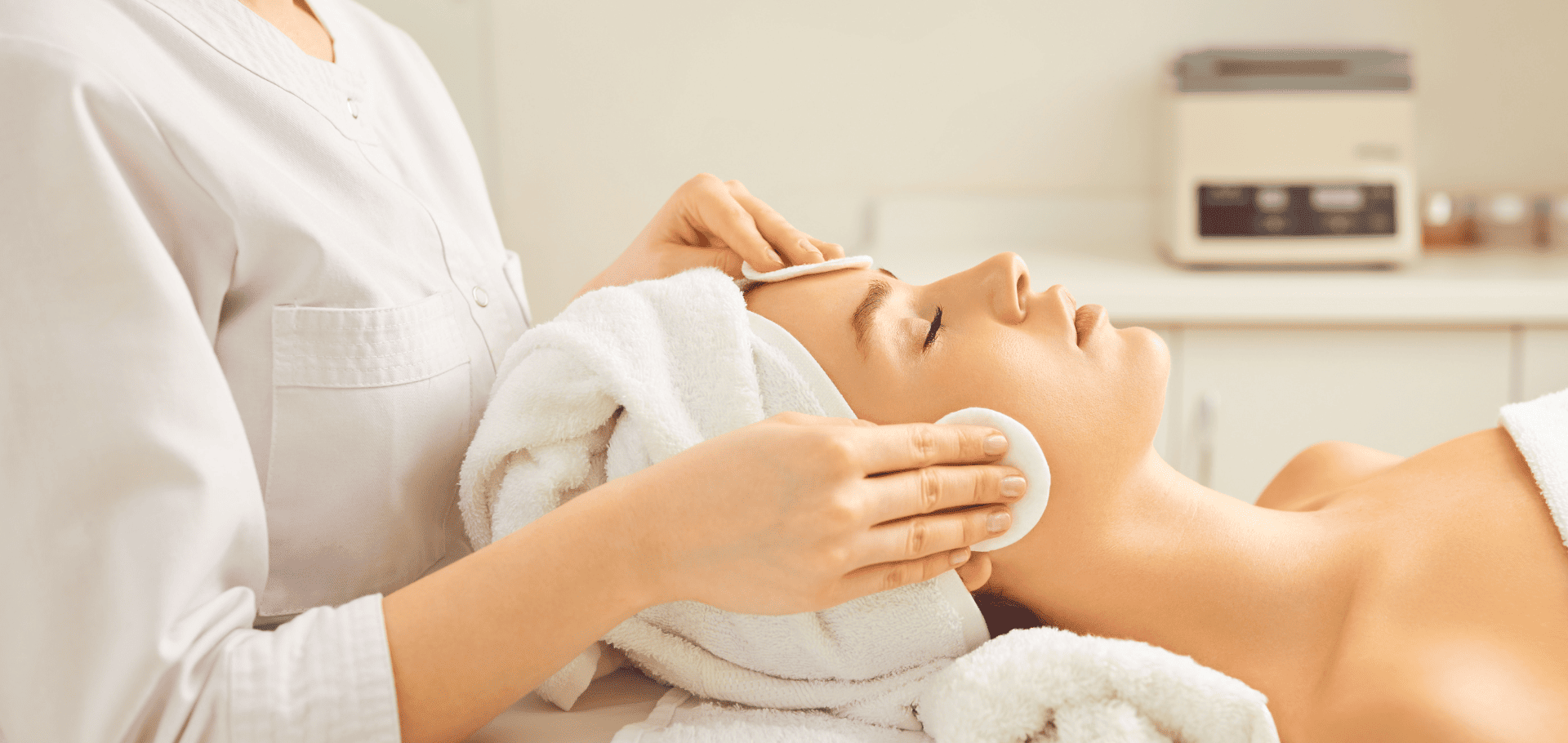 Leząca kobieta z ładną cerą i ręcznikiem na głowie, ładną cerą i zamkniętymi oczami podczas gdy kosmetolożka stojąca za nią przemywa jej twarz wacikiem