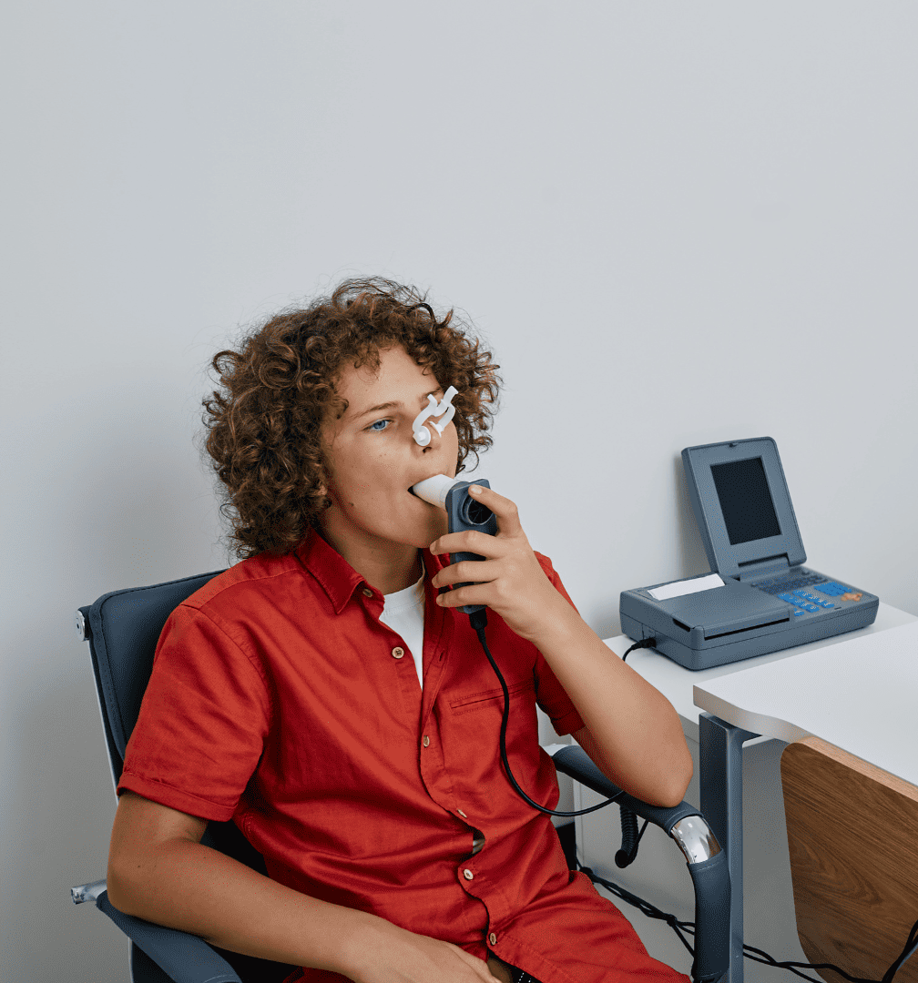 Siedzący chłopiec w czerwonej bluzce, który trzyma ustnik w buzi oraz ma medyczny klips na nosie podczas wykonywania badania spirometrycznego.