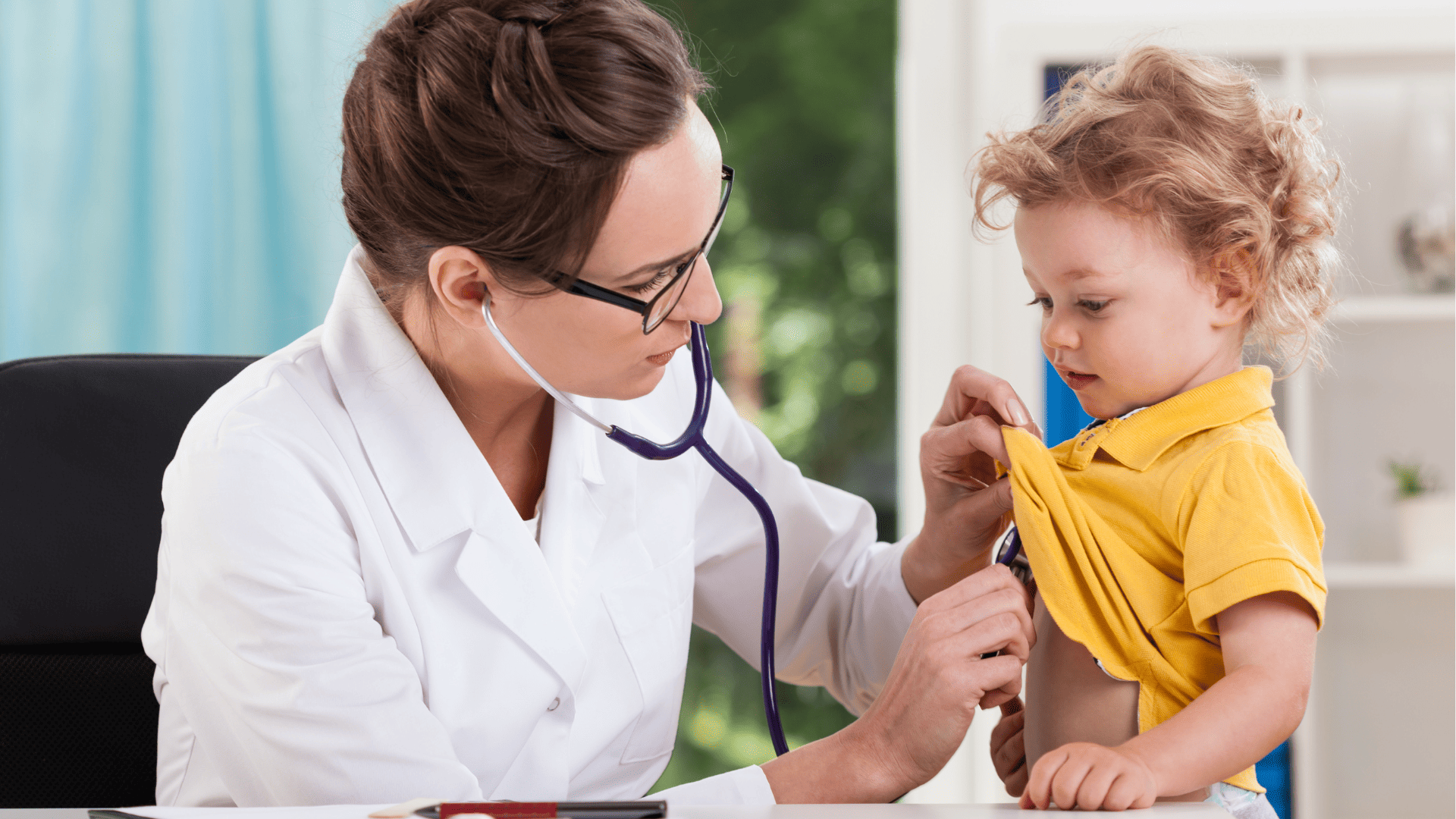 Doktor w białym fartuchu i okularach osłuchująca małe dziecko w żółtej koszulce, w wieku ok. 4 lat za pomocą stetoskopu