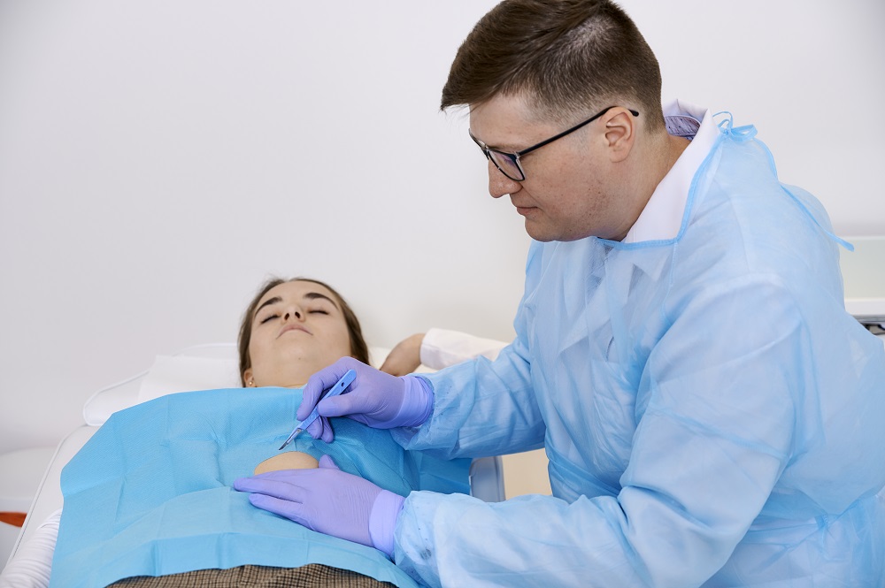 Spokojnie uśmiechnięty chirurg lek. Aleksander Husak usuwa zmianę skórną nachylając się nad pacjentką, która ma na sobie chustę chirurgiczną