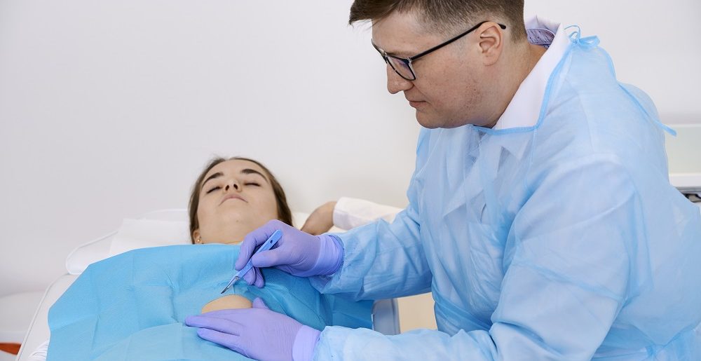 Spokojnie uśmiechnięty chirurg lek. Aleksander Husak usuwa zmianę skórną nachylając się nad pacjentką, która ma na sobie chustę chirurgiczną